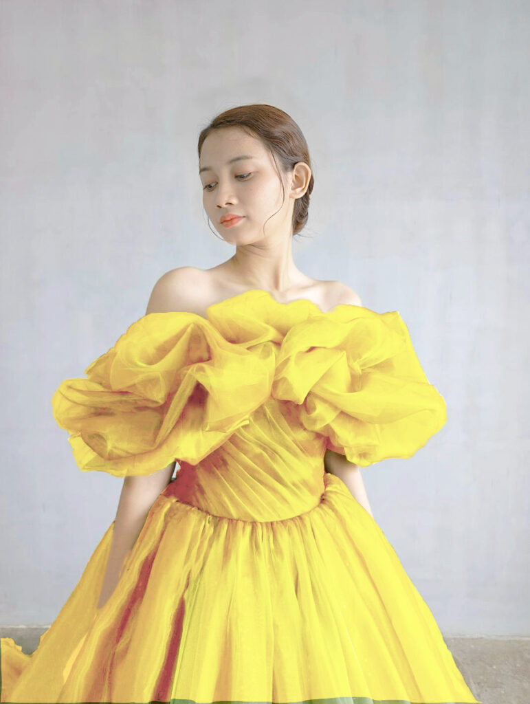 ST-100（イエロー） | 光をまとうような透明感と、空気をはらんだような柔らかさが魅力のボリュームドレス。ふんわりと立体的なオーガンジースリーブが顔まわりを華やかに演出し、まるで花びらをまとったような印象に。スリーブは取り外し可能で、表情の変化を楽しめます。今回のモデルは、特別仕様のイエローカラーでオーダーされた一点。柔らかなレモンイエローが肌を明るく引き立て、晴れやかなウェディングシーンにぴったりのハッピーオーラを纏います。陽の光に溶け込むような軽やかなトレーンと、ドレープの重なりが生み出す立体感が、見る人の視線を惹きつける一着です。
