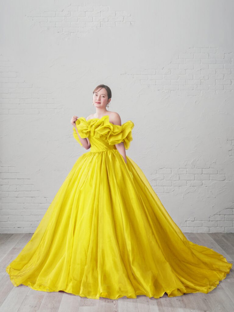 ST-100（イエロー） | 光をまとうような透明感と、空気をはらんだような柔らかさが魅力のボリュームドレス。ふんわりと立体的なオーガンジースリーブが顔まわりを華やかに演出し、まるで花びらをまとったような印象に。スリーブは取り外し可能で、表情の変化を楽しめます。今回のモデルは、特別仕様のイエローカラーでオーダーされた一点。柔らかなレモンイエローが肌を明るく引き立て、晴れやかなウェディングシーンにぴったりのハッピーオーラを纏います。陽の光に溶け込むような軽やかなトレーンと、ドレープの重なりが生み出す立体感が、見る人の視線を惹きつける一着です。