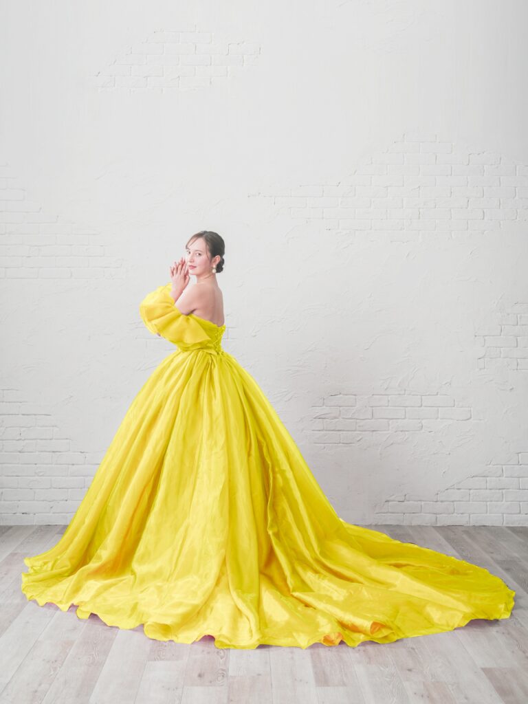 ST-100（イエロー） | 光をまとうような透明感と、空気をはらんだような柔らかさが魅力のボリュームドレス。ふんわりと立体的なオーガンジースリーブが顔まわりを華やかに演出し、まるで花びらをまとったような印象に。スリーブは取り外し可能で、表情の変化を楽しめます。今回のモデルは、特別仕様のイエローカラーでオーダーされた一点。柔らかなレモンイエローが肌を明るく引き立て、晴れやかなウェディングシーンにぴったりのハッピーオーラを纏います。陽の光に溶け込むような軽やかなトレーンと、ドレープの重なりが生み出す立体感が、見る人の視線を惹きつける一着です。