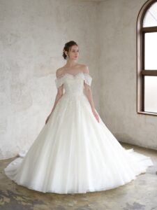 Fi-34（アイボリー）| すっきりと美しいシルエットに、上品な煌めきが洗練された印象のウエディングドレスです。ドレス全面に編み目模様のチュールにグリッターを施した煌めきの素材を重ねて、輝きだけでなくハイクラスな質感を感じさせます。また、レースは繊細なリーフ柄に細やかなビーディングをケープから流れるように施しました。肩周りを透かせることで軽やかに、ハートネックラインが女性らしい優しい表情を演出します。