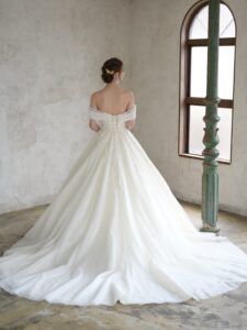 Fi-34（アイボリー）| すっきりと美しいシルエットに、上品な煌めきが洗練された印象のウエディングドレスです。ドレス全面に編み目模様のチュールにグリッターを施した煌めきの素材を重ねて、輝きだけでなくハイクラスな質感を感じさせます。また、レースは繊細なリーフ柄に細やかなビーディングをケープから流れるように施しました。肩周りを透かせることで軽やかに、ハートネックラインが女性らしい優しい表情を演出します。