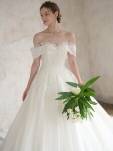 Fi-34（アイボリー）| すっきりと美しいシルエットに、上品な煌めきが洗練された印象のウエディングドレスです。ドレス全面に編み目模様のチュールにグリッターを施した煌めきの素材を重ねて、輝きだけでなくハイクラスな質感を感じさせます。また、レースは繊細なリーフ柄に細やかなビーディングをケープから流れるように施しました。肩周りを透かせることで軽やかに、ハートネックラインが女性らしい優しい表情を演出します。