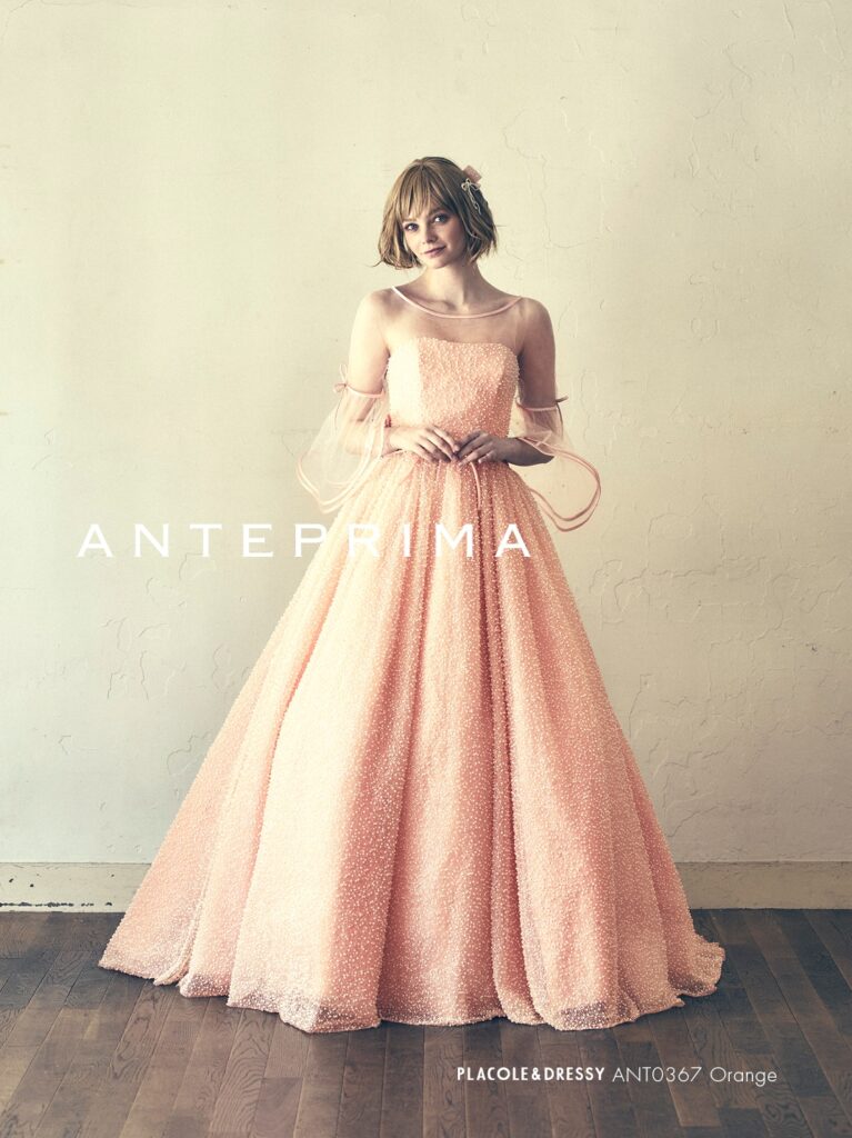 ANT0367（アイボリー/オレンジ） | ジュエルパールグリッターの輝きを全身にまとい、上品さと可憐さをあわせ持つ“現代のプリンセス”を表現。新素材“エアーファブリックオーガン”が、ふんわりと空気を含んだような軽やかなドレープを描きます。ピーチオレンジのやさしい色合いが肌にやわらかく映え、見る人にぬくもりと幸せを届けるような一着です。