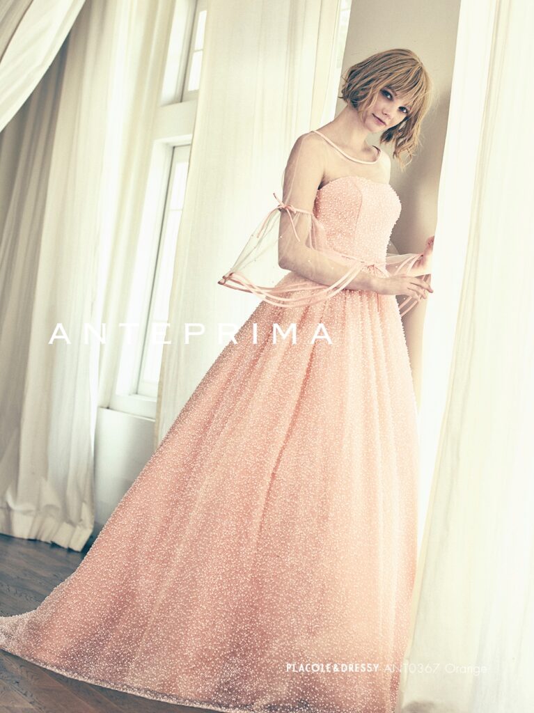 ANT0367（アイボリー/オレンジ） | ジュエルパールグリッターの輝きを全身にまとい、上品さと可憐さをあわせ持つ“現代のプリンセス”を表現。新素材“エアーファブリックオーガン”が、ふんわりと空気を含んだような軽やかなドレープを描きます。ピーチオレンジのやさしい色合いが肌にやわらかく映え、見る人にぬくもりと幸せを届けるような一着です。