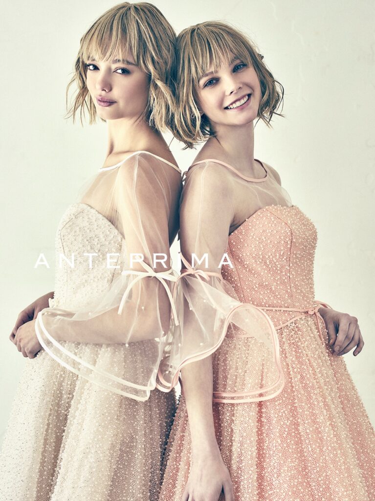 ANT0367（アイボリー/オレンジ） | ジュエルパールグリッターの輝きを全身にまとい、上品さと可憐さをあわせ持つ“現代のプリンセス”を表現。新素材“エアーファブリックオーガン”が、ふんわりと空気を含んだような軽やかなドレープを描きます。ピーチオレンジのやさしい色合いが肌にやわらかく映え、見る人にぬくもりと幸せを届けるような一着です。