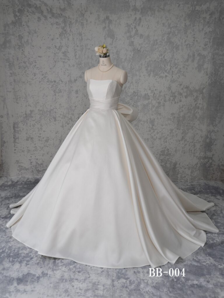 BB-004 | 上品な光沢を放つミカド素材を全面に用いたシンプルなマリエ。幾重にも重ねられてボリューム感を表現したスカートシルエット、ウェストラインにはアシメトリーにタックを取ったサッシュベルト、足長効果を生みます。バックにはアシメトリーの大きなリボンが、スカート裾まで続くクルミボタンと相俟って、シンプルな中に印象的なバックデザインに。