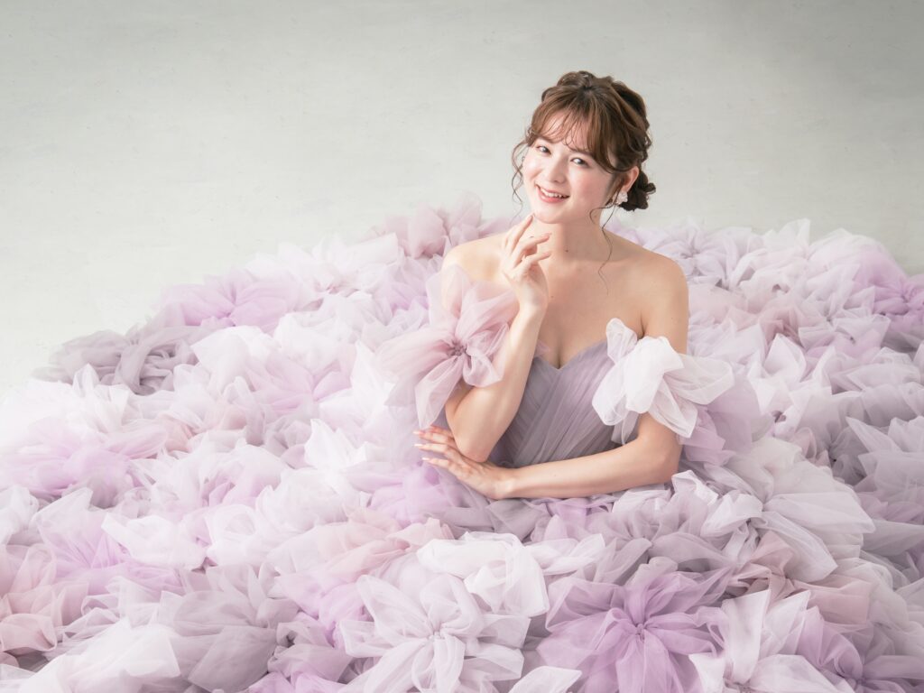 LQ-027（ラベンダー） | 柔らかいピンク・ラベンダーの色を複数混ぜ合わせてひとつずつふんわりと、立体的に形成したハンドメイドフラワーをそのまま身に纏うようなフォトジェニックなドレス。花びらを一枚一枚つまんでふんわり見せる工程や、お花中心のクリスタルをチュールでくるむ技法などよく見るととっても繊細な手仕事でできているのがわかります。