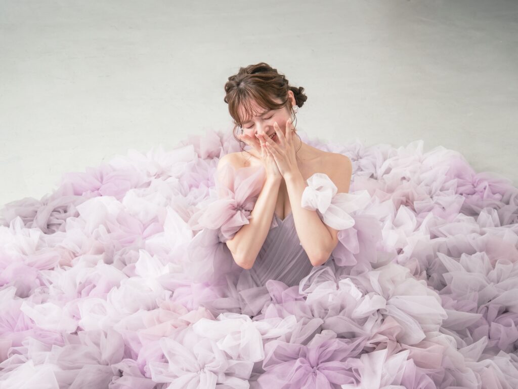 LQ-027(ラベンダー) | 柔らかいピンク・ラベンダーの色を複数混ぜ合わせてひとつずつふんわりと、立体的に形成したハンドメイドフラワーをそのまま身に纏うようなフォトジェニックなドレス。花びらを一枚一枚つまんでふんわり見せる工程や、お花中心のクリスタルをチュールでくるむ技法などよく見るととっても繊細な手仕事でできているのがわかります。