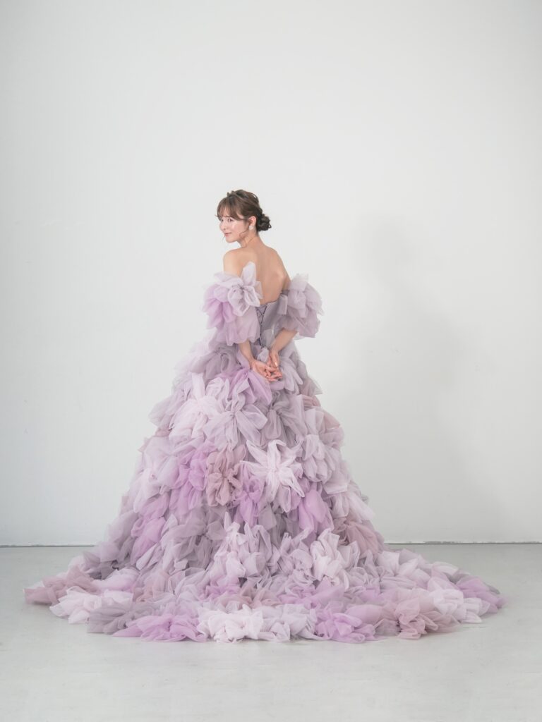 LQ-027（ラベンダー） | 柔らかいピンク・ラベンダーの色を複数混ぜ合わせてひとつずつふんわりと、立体的に形成したハンドメイドフラワーをそのまま身に纏うようなフォトジェニックなドレス。花びらを一枚一枚つまんでふんわり見せる工程や、お花中心のクリスタルをチュールでくるむ技法などよく見るととっても繊細な手仕事でできているのがわかります。