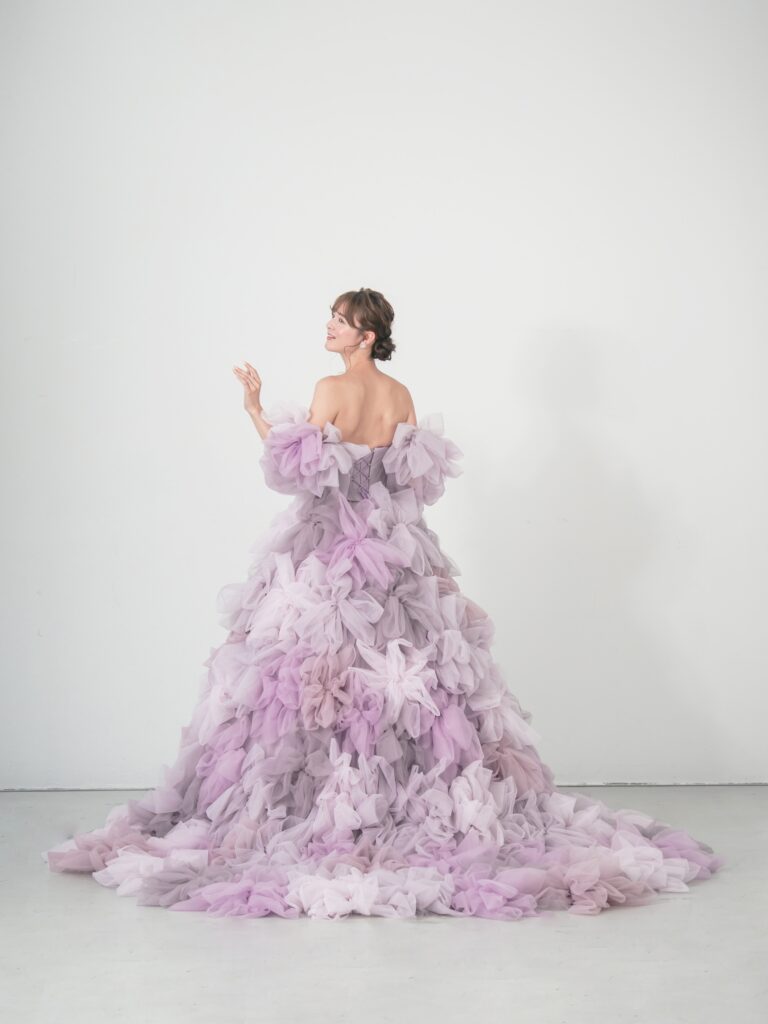 LQ-027（ラベンダー） | 柔らかいピンク・ラベンダーの色を複数混ぜ合わせてひとつずつふんわりと、立体的に形成したハンドメイドフラワーをそのまま身に纏うようなフォトジェニックなドレス。花びらを一枚一枚つまんでふんわり見せる工程や、お花中心のクリスタルをチュールでくるむ技法などよく見るととっても繊細な手仕事でできているのがわかります。