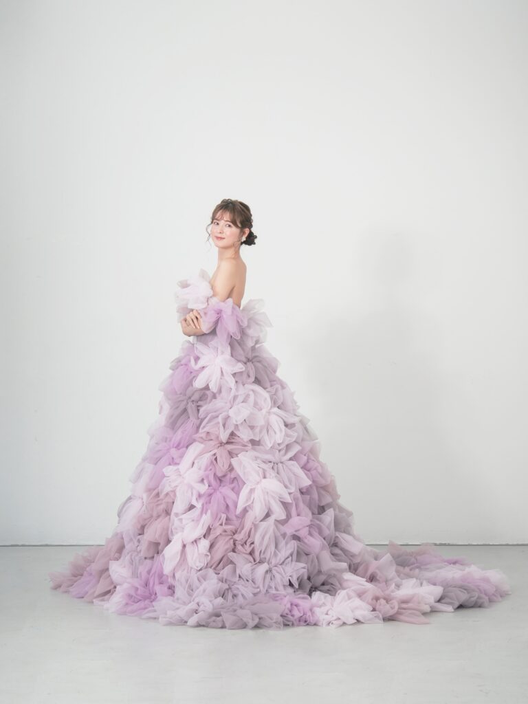 LQ-027（ラベンダー） | 柔らかいピンク・ラベンダーの色を複数混ぜ合わせてひとつずつふんわりと、立体的に形成したハンドメイドフラワーをそのまま身に纏うようなフォトジェニックなドレス。花びらを一枚一枚つまんでふんわり見せる工程や、お花中心のクリスタルをチュールでくるむ技法などよく見るととっても繊細な手仕事でできているのがわかります。