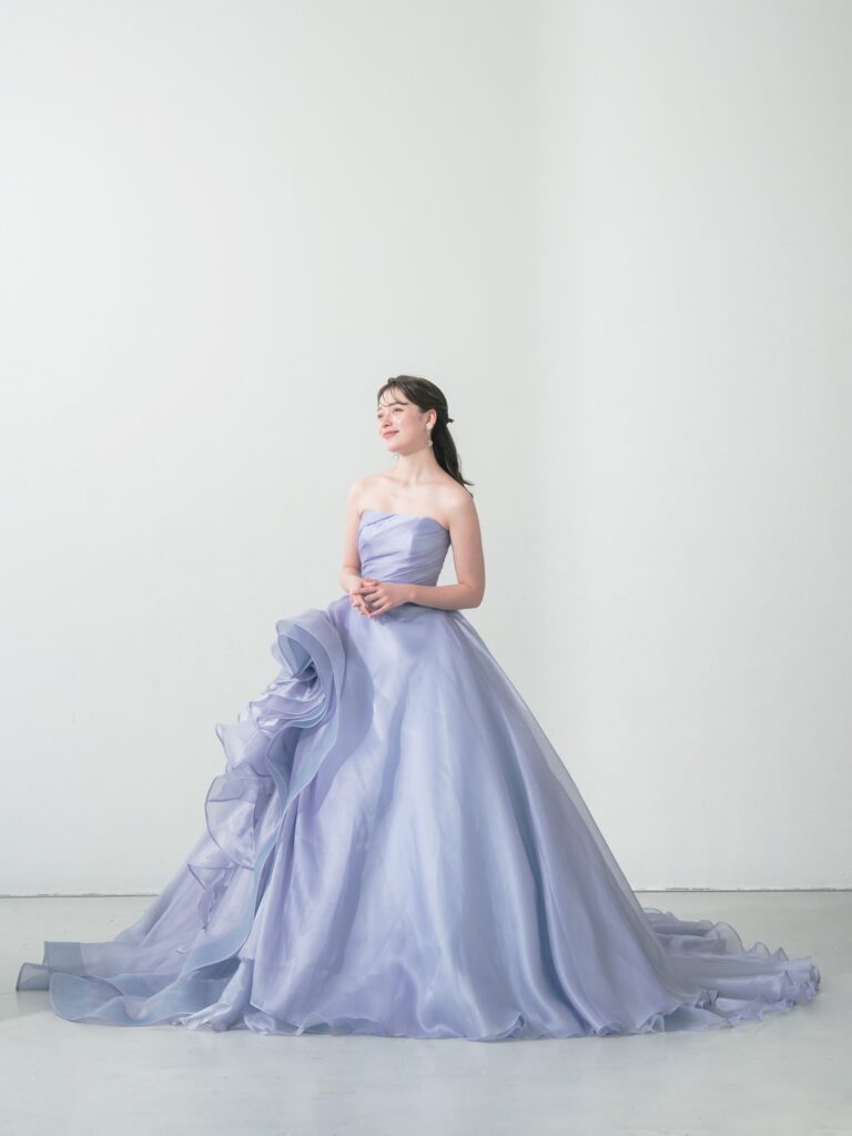ST-111 | やわらかなウィスティリアカラーが空気まで優しく染める一着。透明感のあるオーガンジーの重なりと、流れるようなボリュームシルエットが、動くたびに表情を変えてくれます。 甘すぎず、上品で、記憶に残るカラードレス。静かな華やかさをまといたい花嫁さまへ。