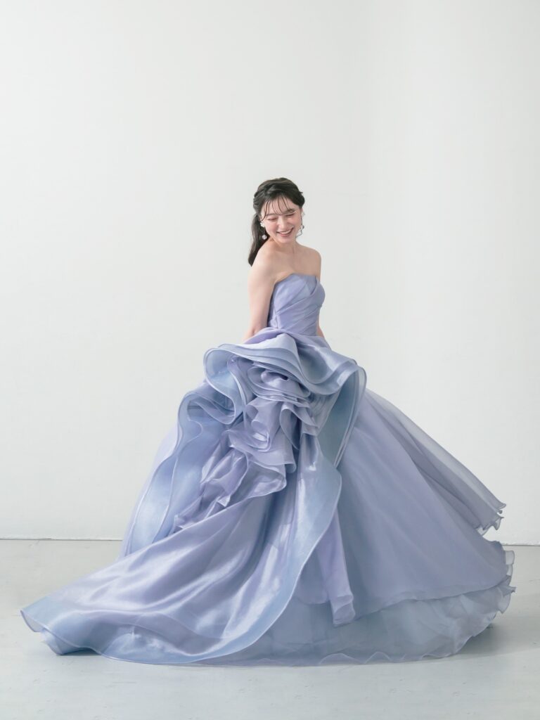 ST-111 | やわらかなウィスティリアカラーが空気まで優しく染める一着。透明感のあるオーガンジーの重なりと、流れるようなボリュームシルエットが、動くたびに表情を変えてくれます。 甘すぎず、上品で、記憶に残るカラードレス。静かな華やかさをまといたい花嫁さまへ。